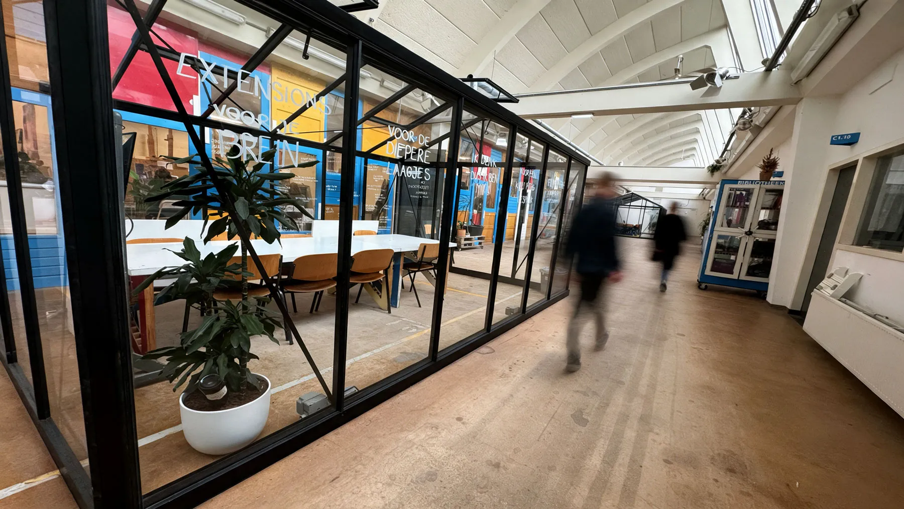 lange gang langs glazen werkruimtes met planten, iemand loopt voorbij